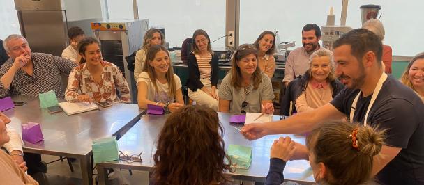 El chef catalán Llorenç Sagarra ubicado en la cabecera de una mesa comparte un producto a una asistente de su taller, ante la risa de las personas participantes