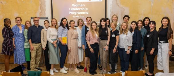 A group of people (mostly women but also a few men) are standing in front of a big screen with the inscription "Women Leadership Programme"