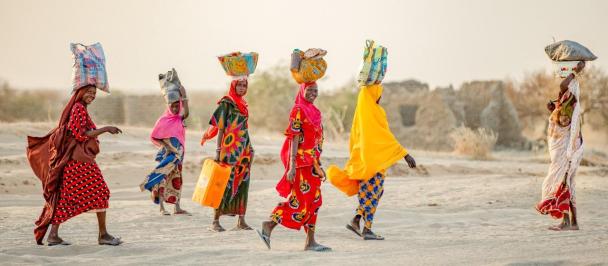 Women in brightly coloured clothing walk across sand carrying water vessels on their heads,