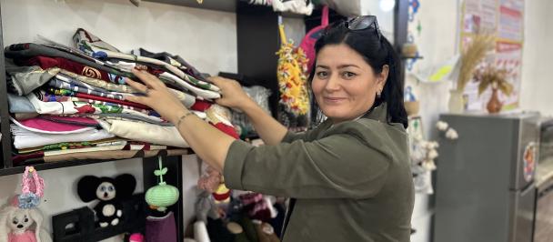 A woman with shoulder-length dark hair and sunglasses perched atop her head smiles at the camera while selecting fabrics from a shelf in a well-organized textile workshop. She is wearing a casual olive-green dress and has a wristwatch on her left arm. The shelf is filled with neatly folded textiles of various patterns and colors. In the foreground, a display features an assortment of colorful handmade crafts, including plush toys and decorative items.