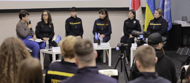 A panel discussion is taking place in a room with a group of women seated in front of an audience. The panelists, some of whom are dressed in black uniforms with yellow trim, are sitting behind a table with name tags and microphones. There are national flags of Ukraine, Japan and the United Nations behind them. In the foreground, the back of the audience is visible, mostly consisting of individuals wearing jackets with "Rescuer" inscription.