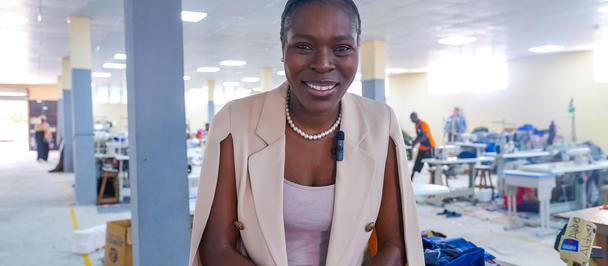 A smiling woman in a beige vest stands in a sewing workshop with various fabrics around.