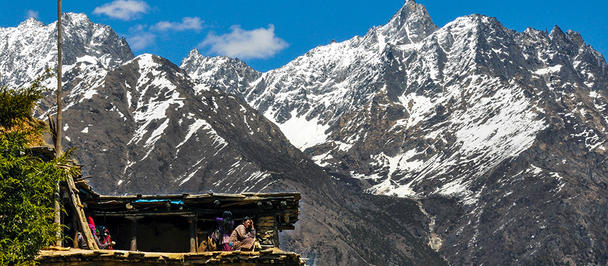 Snow-capped mountains loom over a rustic wooden structure with people sitting outside.