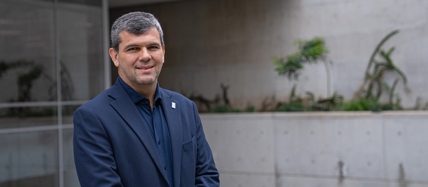 Smiling man in a dark blazer stands outdoors near a modern building with greenery.