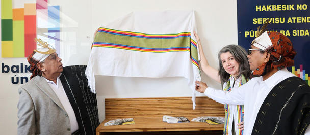 Three people, dressed in traditional attire, unveil a colorful textile in a ceremonial setting.