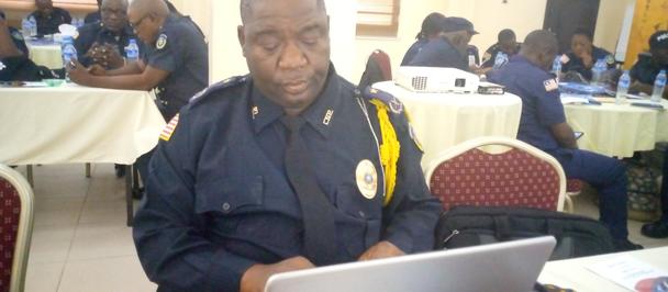 A focused man in a police uniform sits at a laptop in a conference room.