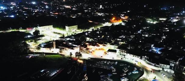 Aerial view of a bustling town at night, illuminated streets, and harbor with boats.