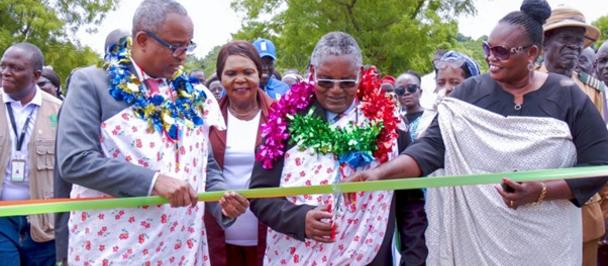 Officials in colorful garlands cut a ribbon at an outdoor ceremony, surrounded by an audience.