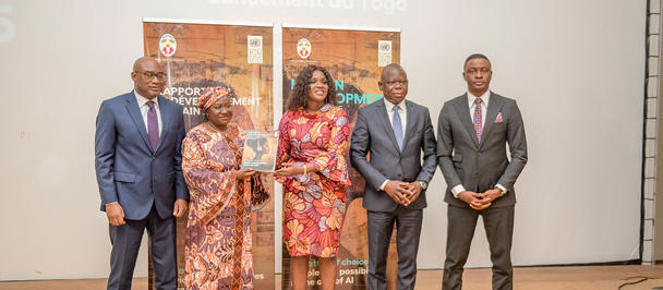 Group photo: six people in formal attire on stage at a conference, with a projection screen behind.