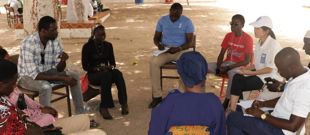 Group of people seated in a circle outdoors in a dusty yard under a tree.