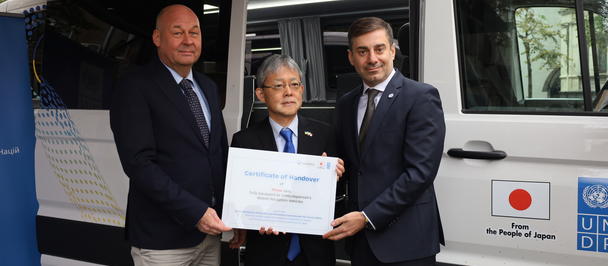 Three people pose with a certificate in front of a logo-covered van displaying a Japan flag.