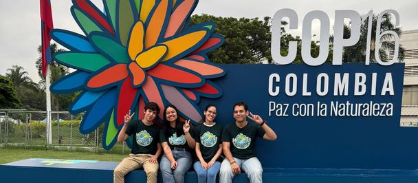 Group of people posing in front of a colorful flower sculpture and COP16 Colombia banner.