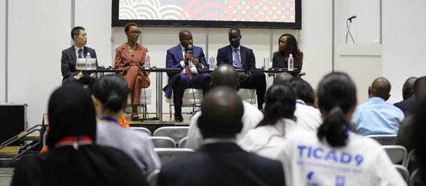 Panel discussion on stage with six speakers; audience seated in a conference room.
