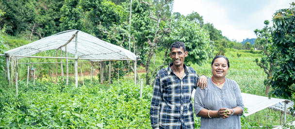 Two smiling people stand amidst lush greenery, with a cultivation structure in the background.