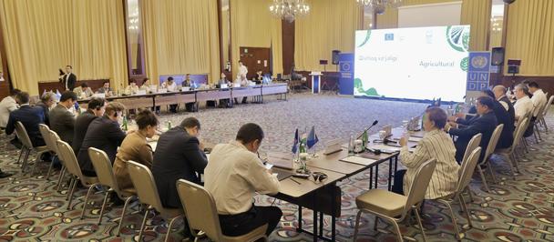 Large conference room with participants seated around U-shaped tables, projection screen at front.