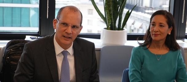 Two people sit at a table in an office, man in suit left, woman in teal right.