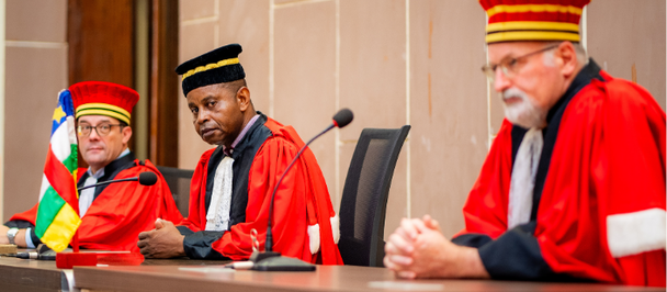 Three judges in red and black ceremonial robes seated at a panel desk.