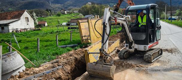 Compact excavator digging a trench along a rural road; worker in safety gear; mountains.