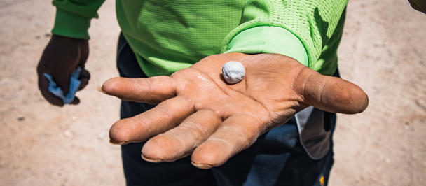 Person in green shirt extends hand toward camera, palm showing a coin; blue item in the other hand.