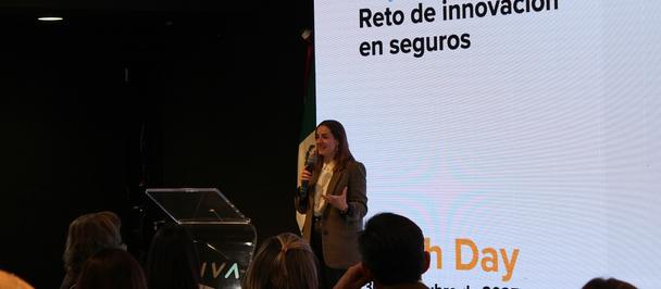 Female presenter on stage at a conference with a Spanish slide behind her, audience in foreground.