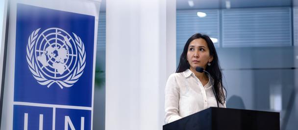 Photograph of a woman speaking at a podium beside a UN banner.