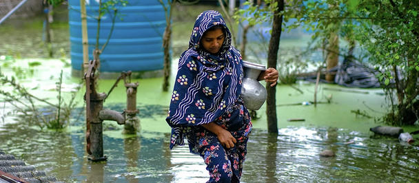 Girl in a blue patterned hijab walks through floodwaters beside trees.
