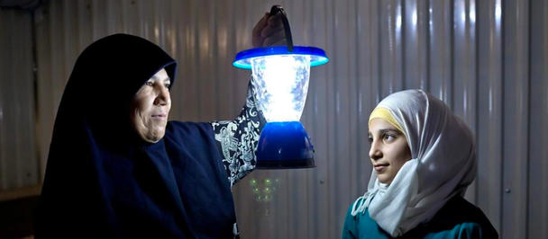 Two people in a dim room hold up lantern glowing with blue light, with metal walls behind them