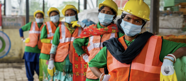 Line of construction workers wearing orange vests, yellow hard hats, and masks at a site.
