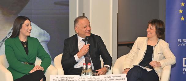 Photograph: three panelists in business attire on stage with a blue EU banner behind.