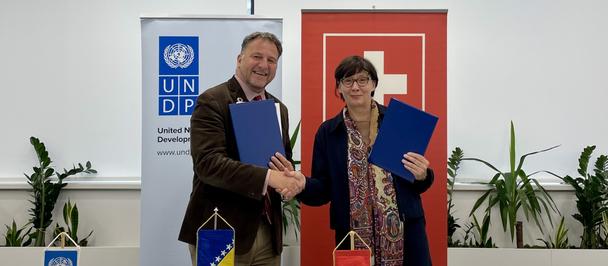 Two people sign documents and shake hands at a signing ceremony; UNDP banner and flags visible.