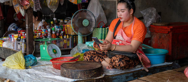 Market stall cook in orange top, preparing meat amid bowls, a fan, and hanging goods.