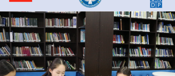 Photograph: three children reading at a library table, shelves behind, flags and logos overhead.