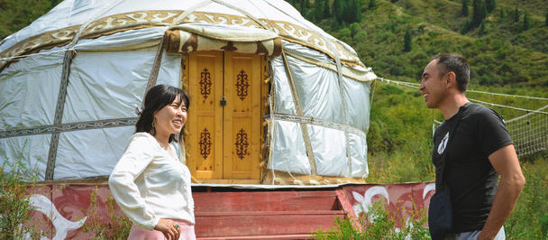 Two people converse beside a white yurt on a red deck in a green hillside landscape.