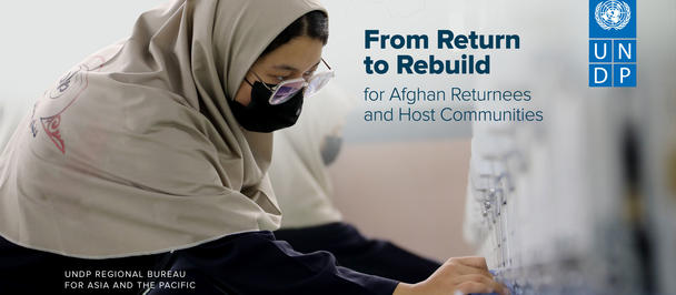 Woman in hijab and mask writes at a desk beside a UNDP banner reading From Return to Rebuild.