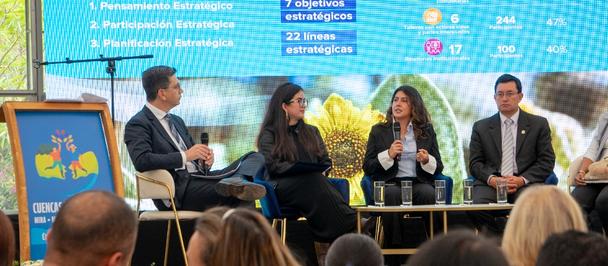 Panelists and moderator on stage at a conference, blue program screen behind, audience in front.