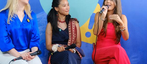 Three women with a blue backdrop; left in blue, middle in dark dress, right in red with mic.