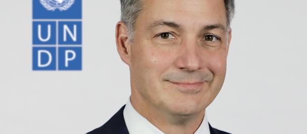 Portrait of a man in a dark suit and red tie, with a blue UNDP logo in the background.