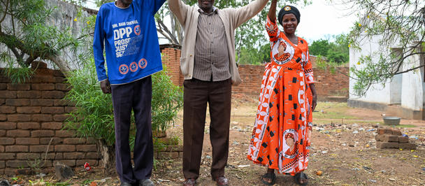 Three people outdoors, standing on dirt, raising hands in celebration; brick wall and trees behind.