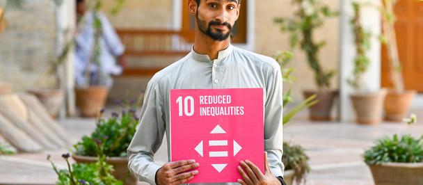 Bearded man in a gray shirt holds a pink square sign with white arrows outdoors.