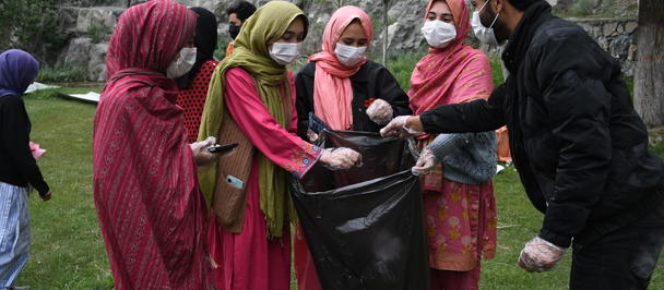 Group of women in colorful hijabs outdoors, a man hands an item to a woman near a large trash bag.