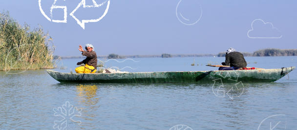 Two people paddling a small green boat on calm blue water; reeds on the shore, with logos at bottom