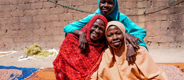 Three women in vibrant traditional attire pose on colorful mats outdoors, brick wall behind.