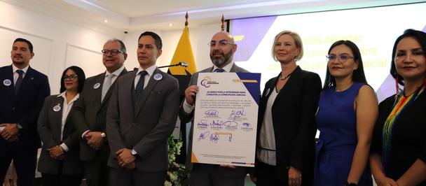 Group of formally dressed people posing with a framed certificate indoors, flags in the background.