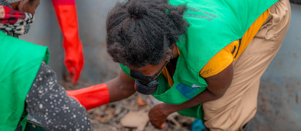 Two people in green safety vests lean over a green dumpster, handling a red strap.