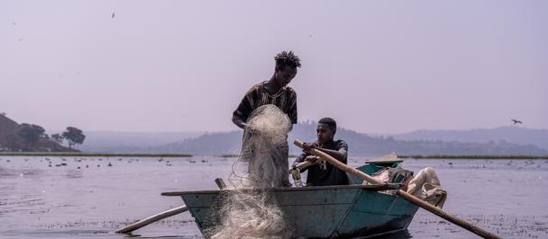 Photograph: man standing at the bow of a blue boat on calm water, splashing water as another person sits