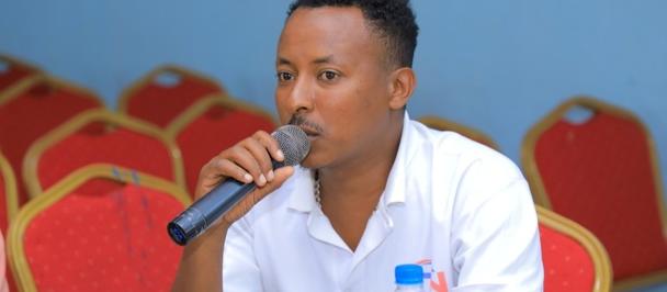 Speaker in white shirt speaks into a microphone, seated with water bottles nearby, red chairs in the background.