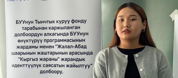 Young woman in a gray cardigan standing beside a banner with logos and text.