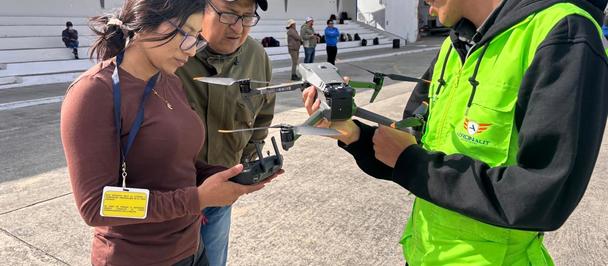 Tres personas al aire libre; un miembro del personal con chaleco neón demuestra el uso de un dron a dos observadores.