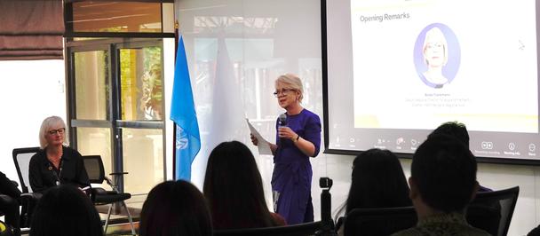 Speaker in purple at a podium presenting to an audience; slide shows portrait, blue flag visible.