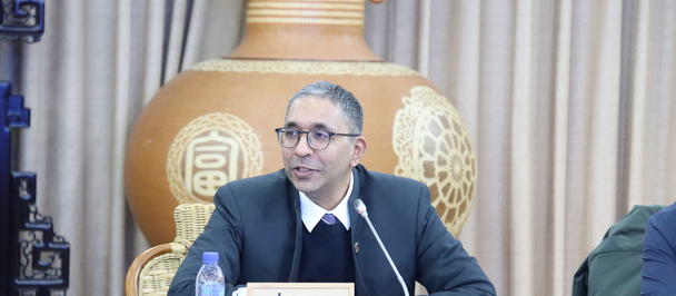 Conference panel with a suited man, blue table, nameplate James George, ornate vase backdrop.